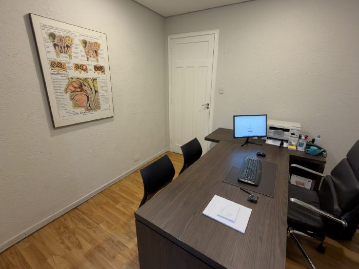 Sala de atendimento com mesa, cadeiras para o paciente e quadro de anatomia do nariz na parede